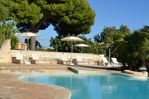 Piscina con sillas de descanso y sombrillas, rodeada de muros de piedra y árboles.