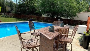 Eine Poolterrasse mit Tisch und Stühlen, umgeben von einem Holenzaun und Bäumen.