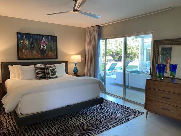 A hotel room with a large bed, a ceiling fan, a painting on the wall, a nightstand with a lamp, and a view of the outdoor pool area. - Oversized Patio, Pool and View! Experience great Indoor/Outdoor living PS style! (Palm Springs)