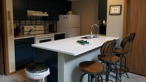 A compact kitchen with a white countertop, black cabinets, a refrigerator, a coffee maker, and a trash can. There are three wooden bar stools and a framed picture on the wall. - 2 Bedroom, 2 bath home in The Galena Territory.  Close to Eagle Ridge Resort (Galena)