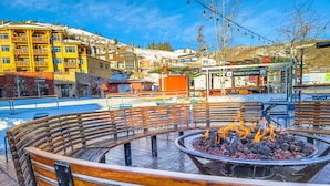 Zona de asientos con bancos de madera y un brasero en el centro, rodeada por una plaza cubierta de nieve y edificios al fondo.