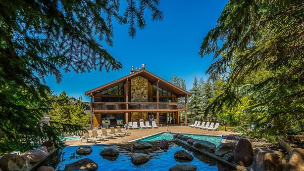 Una cabaña con piscina rodeada de rocas, una terraza con sillones de descanso y un fondo de bosque.