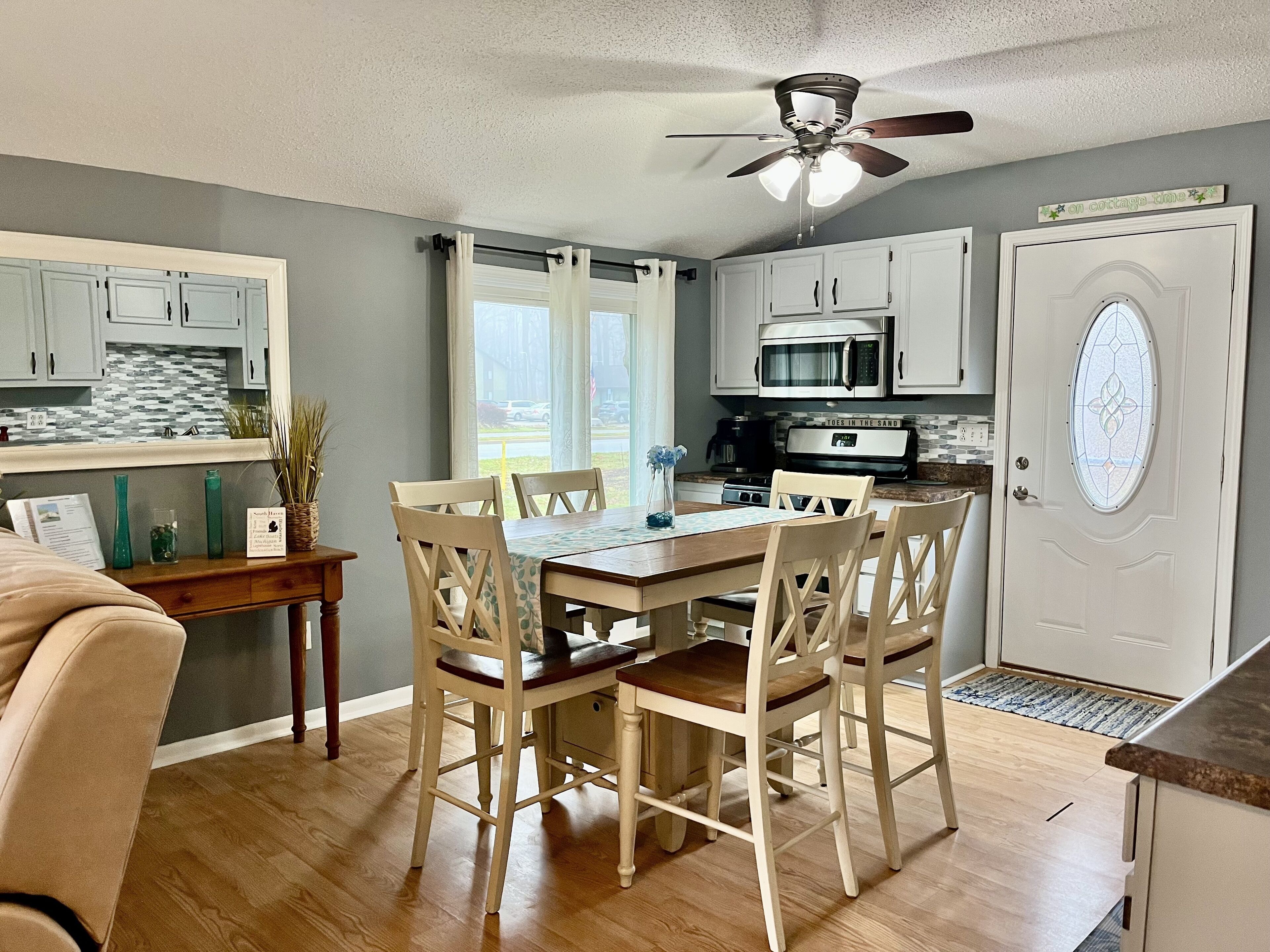 A dining area with a wooden table and chairs, a ceiling fan, and a kitchen with white cabinets and appliances.