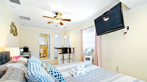 Una habitación de hotel con cama, televisión, ventilador de techo, escritorio con silla y baño visible a través de una puerta abierta.
