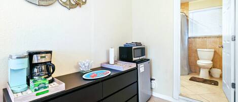 A compact kitchenette with a coffee maker, microwave, and small refrigerator. A decorative wall piece and a door to a bathroom are also visible.