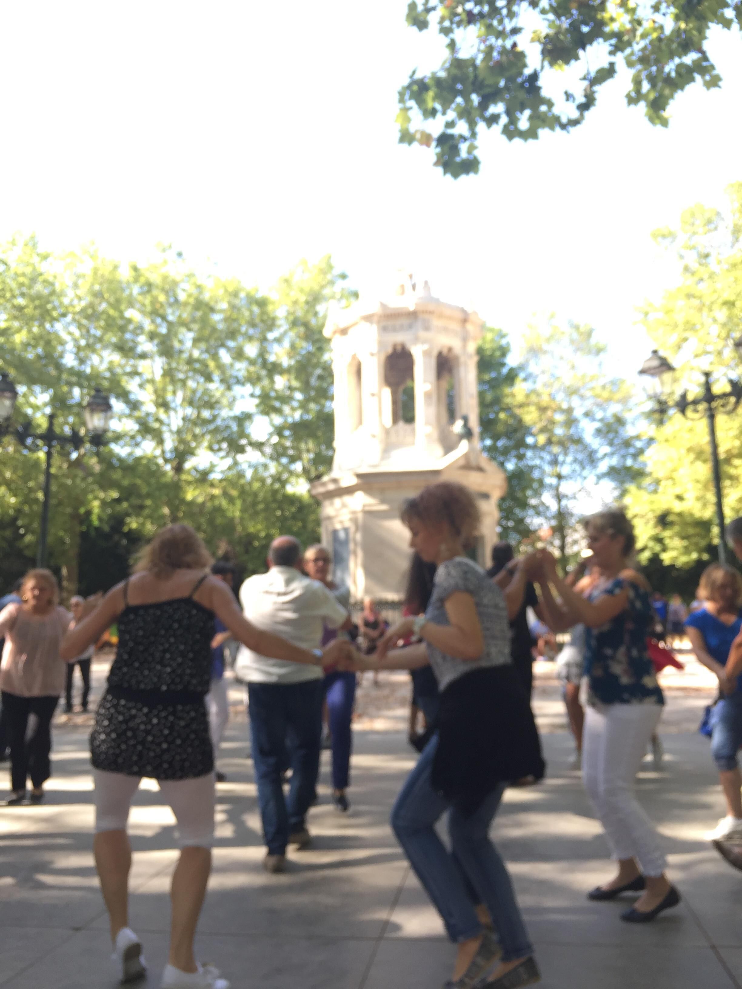 Un grupo de personas bailando en una plaza pública con un monumento histórico al fondo.