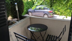 Un petit balcon extérieur avec deux chaises pliantes et une table, une voiture garée dans la rue et de la verdure en arrière-plan.