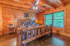 A wooden cabin room with a bed, a ceiling fan, a window, and a small table with a lamp. - True Log Cabin, Hot tub, Fire Pit, Easy access and close to downtown. (Gatlinburg)