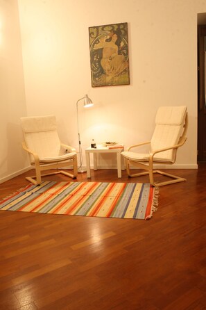 Ein Zimmer mit zwei Holzstühlen, einem kleinen Tisch, einer Lampe und einem farbenfrohen Streifen Teppich auf Holzboden.