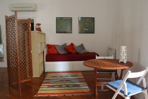 Ein gemütliches Zimmer mit einem Holztisch, einem weißen Stuhl, einer Couch mit roten und grauen Kissen, einem gemusterten Teppich und zwei gerahmten Kunstwerken an der Wand.