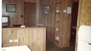A room with wooden paneling, a small kitchenette, a dining area with a table and chairs, and a door leading to another room.