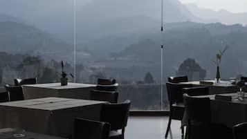 A restaurant with tables and chairs, a potted plant, and a view of mountains.