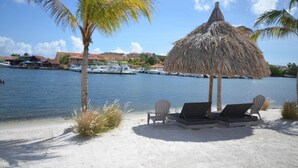 A beach with lounge chairs, a thatched umbrella, and a view of boats and buildings across the water.
