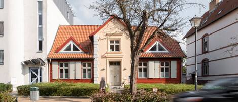 A two-story house with a red-tiled roof, a central entrance, and a small garden area.