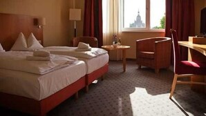 Et hotelværelse med to senge, et skrivebord, en stol, en lampe og et vindue med udsigt til en bygning.