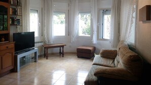 Una sala de estar con televisión, un mueble de madera, un sofá, una mesa y dos ventanas con cortinas.