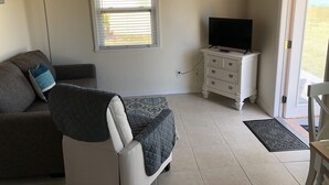 A living room with a sofa, a television on a dresser, and a window with blinds. - Private Beach Front Cottage- unit 1 (Fort Pierce)