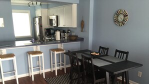 A kitchen with white cabinets, a microwave, and a coffee maker. There is a dining table with four chairs and a black and white checkered rug. - Oceanfront Condo with Fantastic Views (Kure Beach)