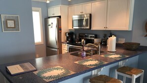 A kitchen with a granite countertop, white cabinets, stainless steel appliances, and bar stools with woven seats. - Oceanfront Condo with Fantastic Views (Kure Beach)