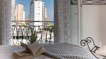Un dormitorio con cama, un libro abierto, colcha estampada, una silla de metal y vistas de edificios altos y una montaña a través de la ventana.