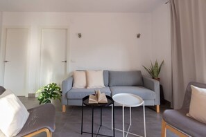 A modern living room with a grey sofa, two side tables, a potted plant, and a door in the background. - Apartamento Paola - Costa CarpeDiem (Calp)