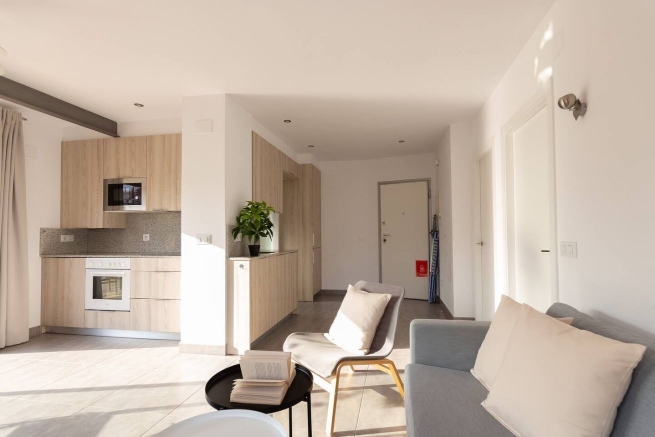 Un salon moderne avec un canapé gris, une petite table sur laquelle repose un livre, et un coin cuisine doté de meubles en bois et d’un four à micro-ondes.