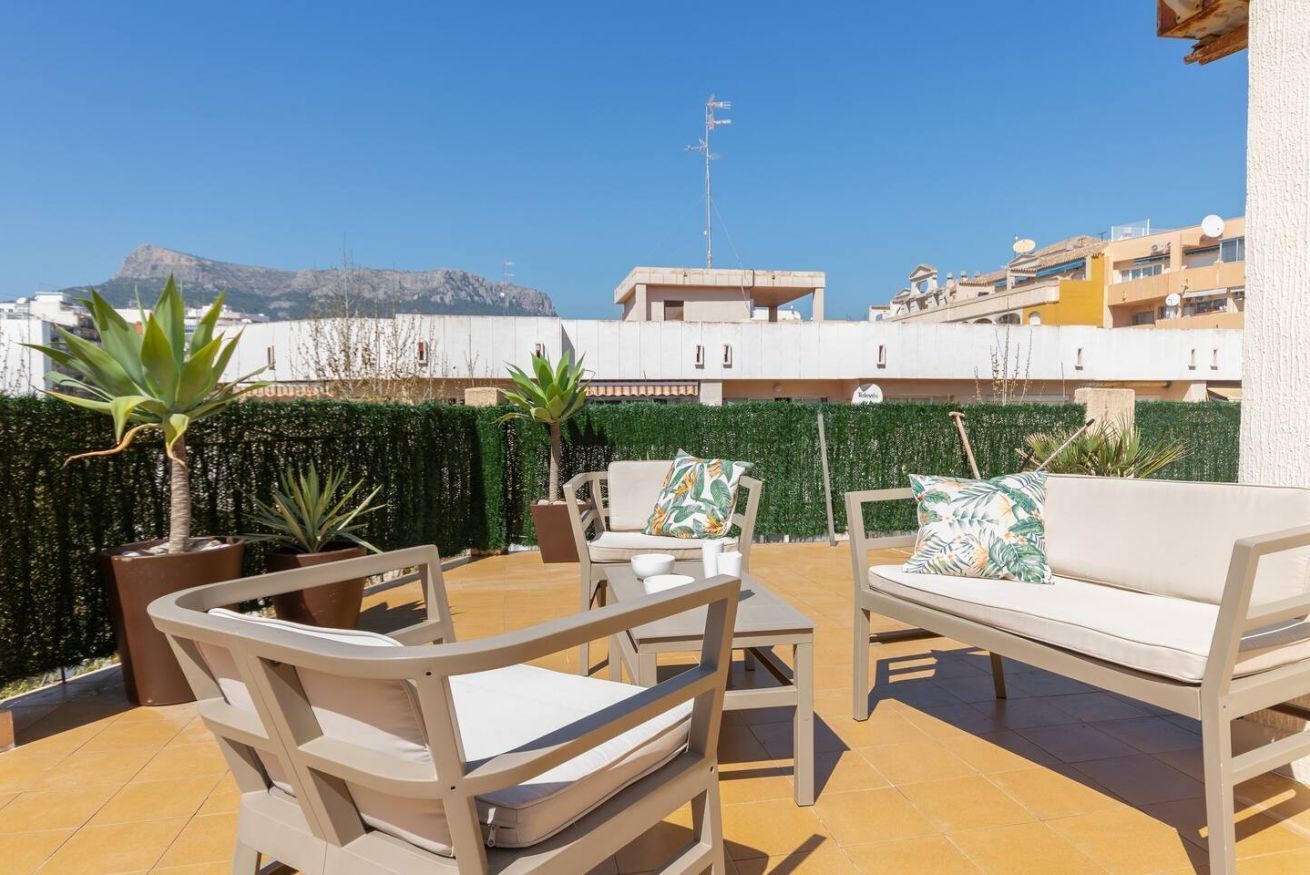 Un patio al aire libre con mobiliario color beige, plantas en macetas y una vista de edificios y una montaña al fondo.