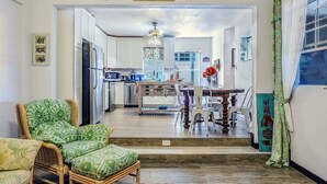Een ruime woonkamer met een eettafel en stoelen, een rieten bank en een keuken op de achtergrond.