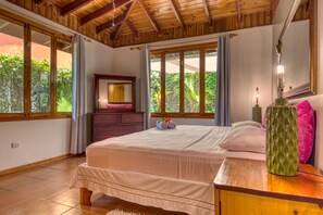 Ein Schlafzimmer mit einem großen Bett, Holzboden und Blick auf Grün hinter großen Fenstern.