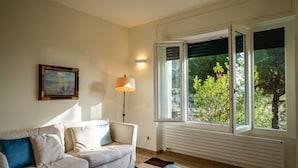 A living room with a white sofa, a glass coffee table, a painting on the wall, and a window with a view of trees. - Villa La Pavona Luxury Collection (Monterosso al Mare)