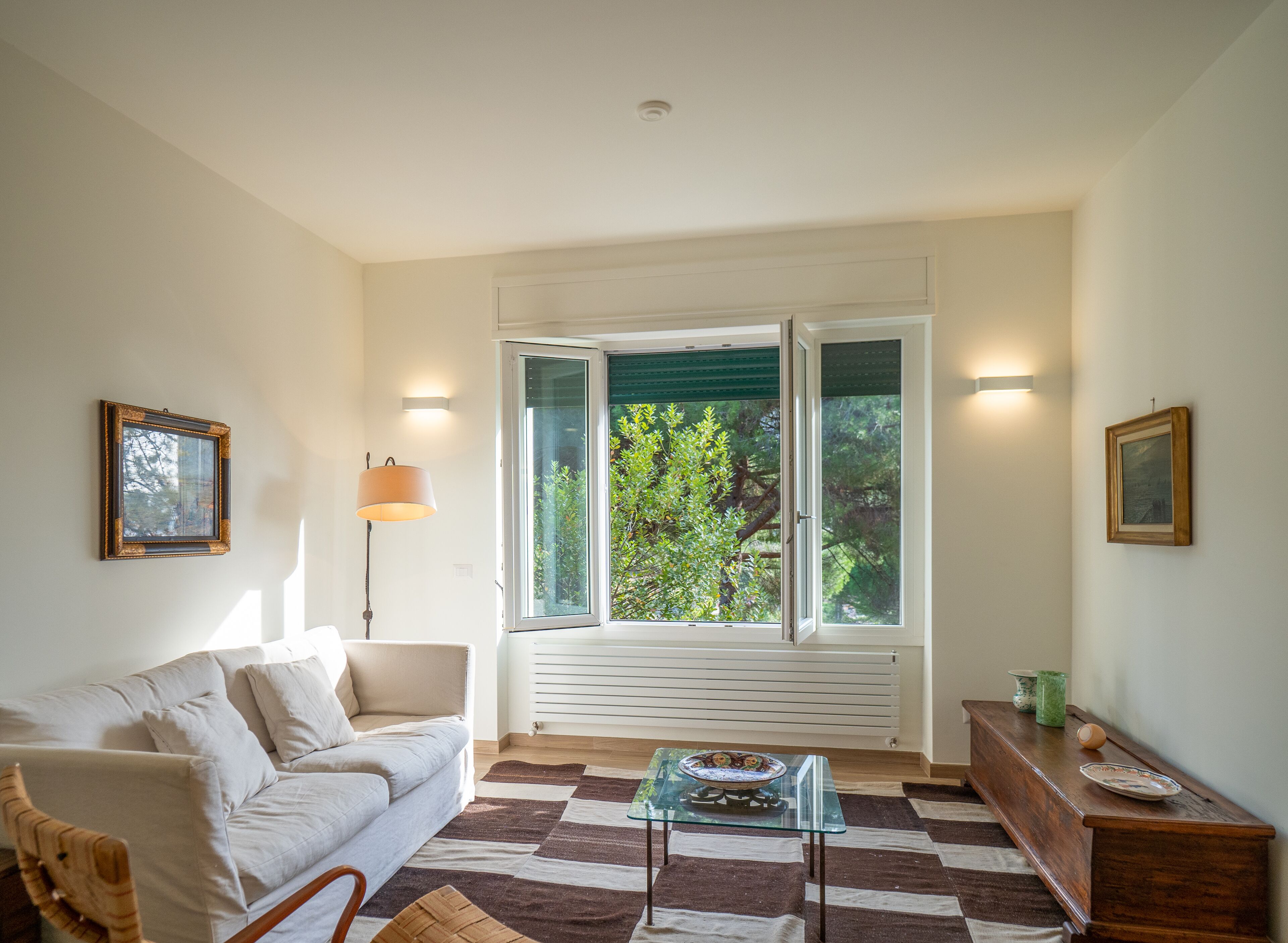 Una sala de estar con un sofá, una mesa de centro de vidrio, una lámpara, una mesita de noche de madera y una ventana con persianas.