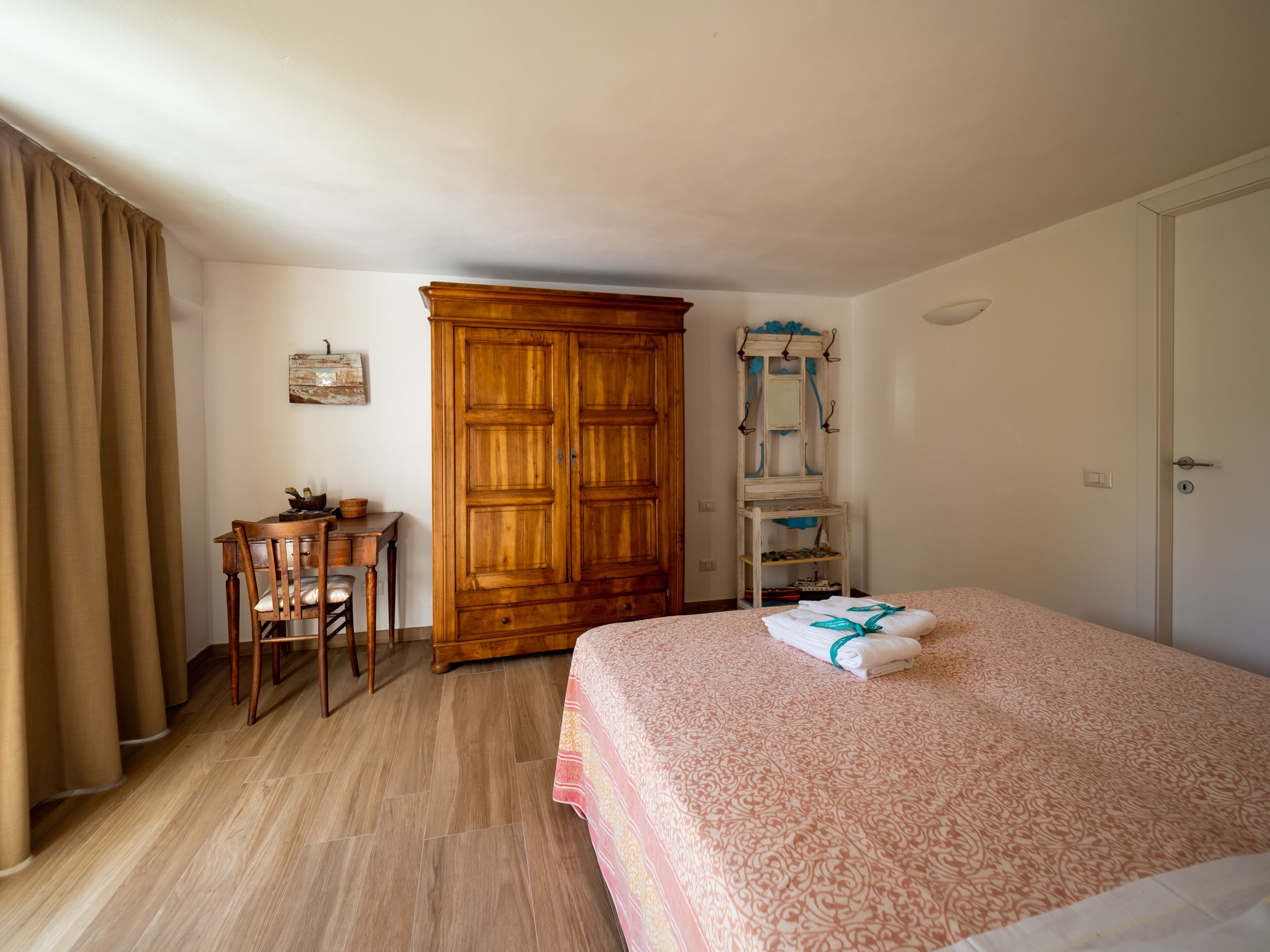 Ein Schlafzimmer mit Holzschrank, Schreibtisch mit Stuhl, Bett mit gemustertem Überwurf und einer Garderobe mit Handtüchern.