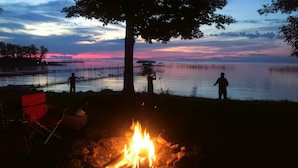 A campfire with a red chair and a person standing by the water.