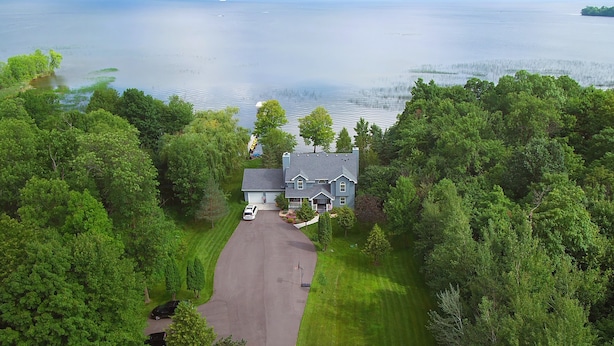 A large house with a driveway and a lawn, surrounded by dense trees and a body of water in the background.