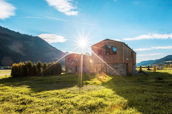 A rustic wooden building with a stone foundation, surrounded by a grassy field and mountains in the background.