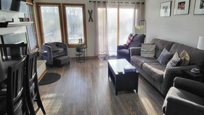 Un salon comprenant un canapé, des fauteuils, une table basse et un téléviseur. Il y a des fenêtres avec des rideaux, une cheminée et des tableaux encadrés accrochés au mur.