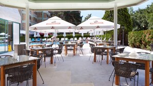 An outdoor patio area with tables and chairs, umbrellas, and a pool in the background. - Aktinia (Nessebar)
