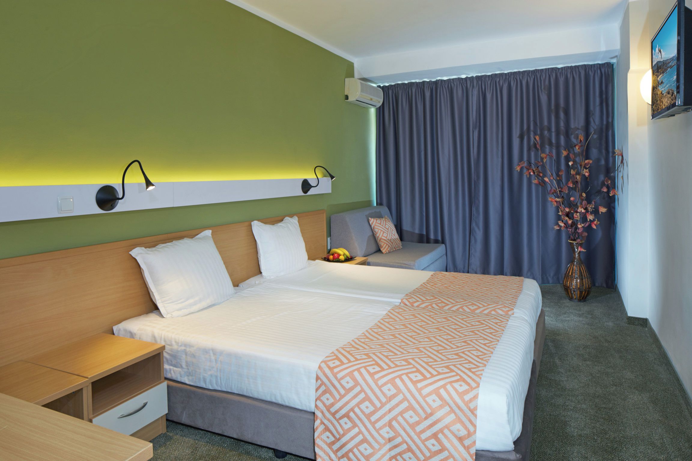 A hotel room with two beds, a desk, a chair, a nightstand, a vase with flowers, and a wall-mounted TV.