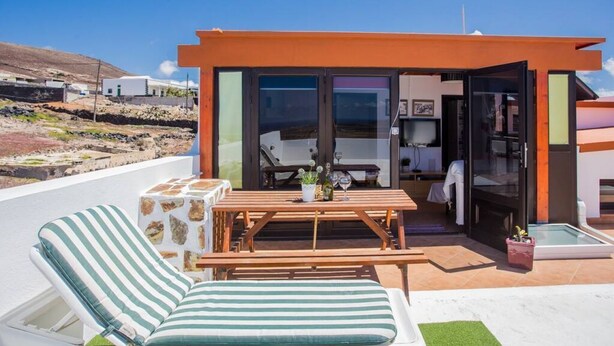 A modern outdoor patio with a wooden table and chairs, a striped lounge chair, and a glass door leading to a room. - Pico Colorado de Soo (Soo)