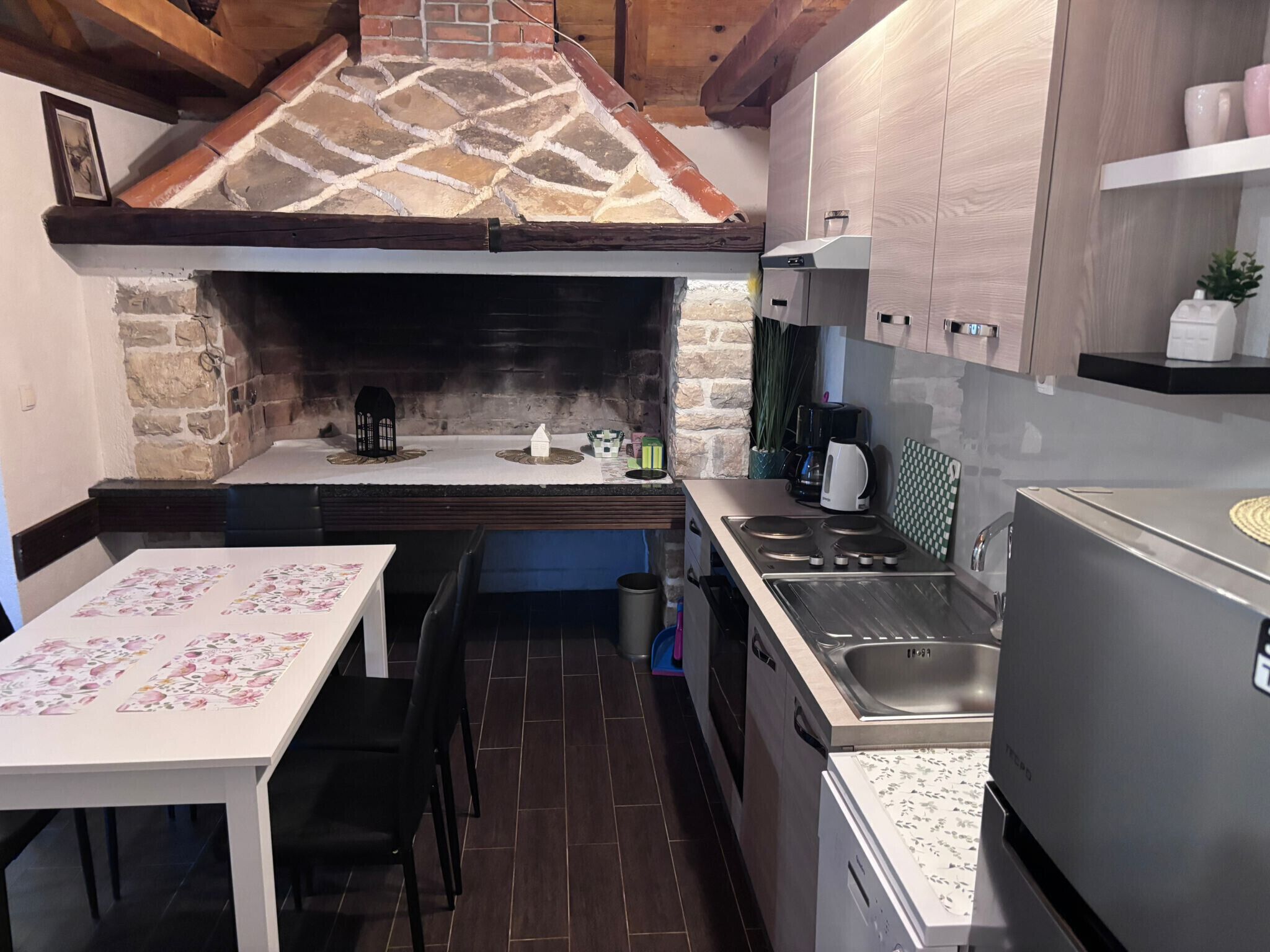 A compact kitchen with a stone fireplace, a dining table with floral tablecloth, a built-in oven, and a refrigerator.