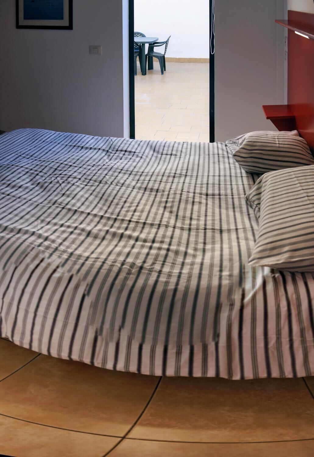 A bed with striped bedding, a tiled floor, and a view of an outdoor area with a table and chairs.