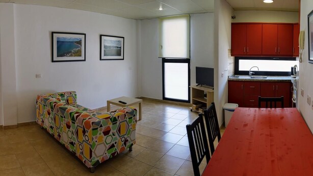 A living room with a colorful sofa, a dining table with chairs, a kitchen area with red cabinets, and two framed pictures on the wall. - Cosy Flat in Playa San Juan with Large Patio (Guía de Isora)