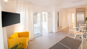 A modern living room with a flat-screen TV, a yellow armchair, a dining area with white chairs, and a glass door leading to an outdoor seating area.