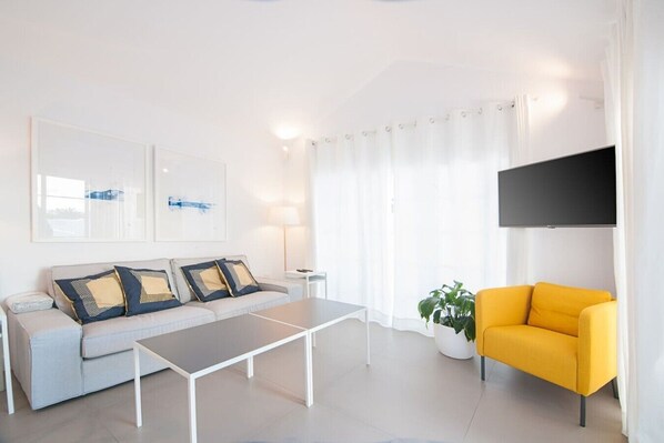 Una sala moderna con un sofá, una mesa de centro y un sillón amarillo. Hay un televisor de pantalla plana montado en la pared y una planta en maceta en el suelo.