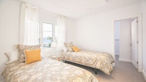 A bedroom with two beds, a window with curtains, and a door leading to another room.