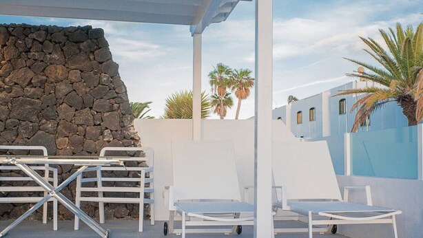 A patio with white chairs and a table, a stone wall, and a blue building with palm trees in the background.