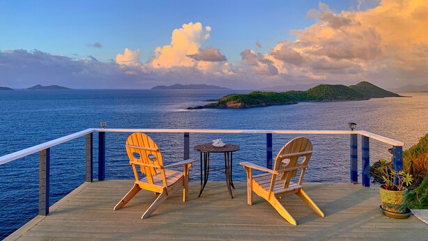 Een houten terras met twee Adirondack stoelen en een kleine tafel, met uitzicht op een rustige zee en verre eilanden.