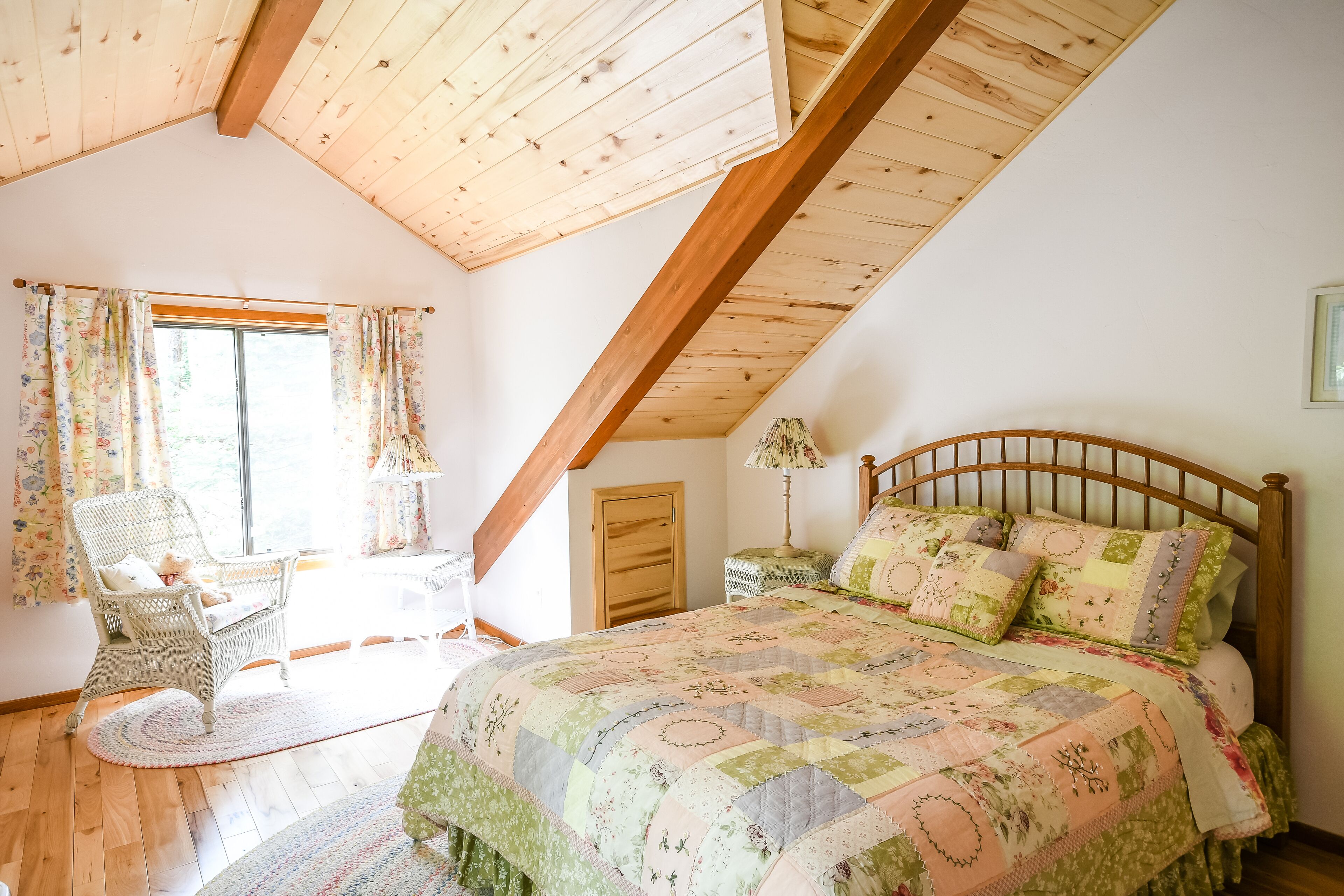 Un dormitorio con techo de madera, una cama grande con ropa de cama floral, una silla de mimbre y una ventana con cortinas.