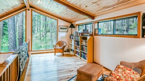 Interior acogedor de una cabaña con pisos de madera, ventanas amplias que ofrecen vistas al bosque, una estantería y un sillón de mimbre.