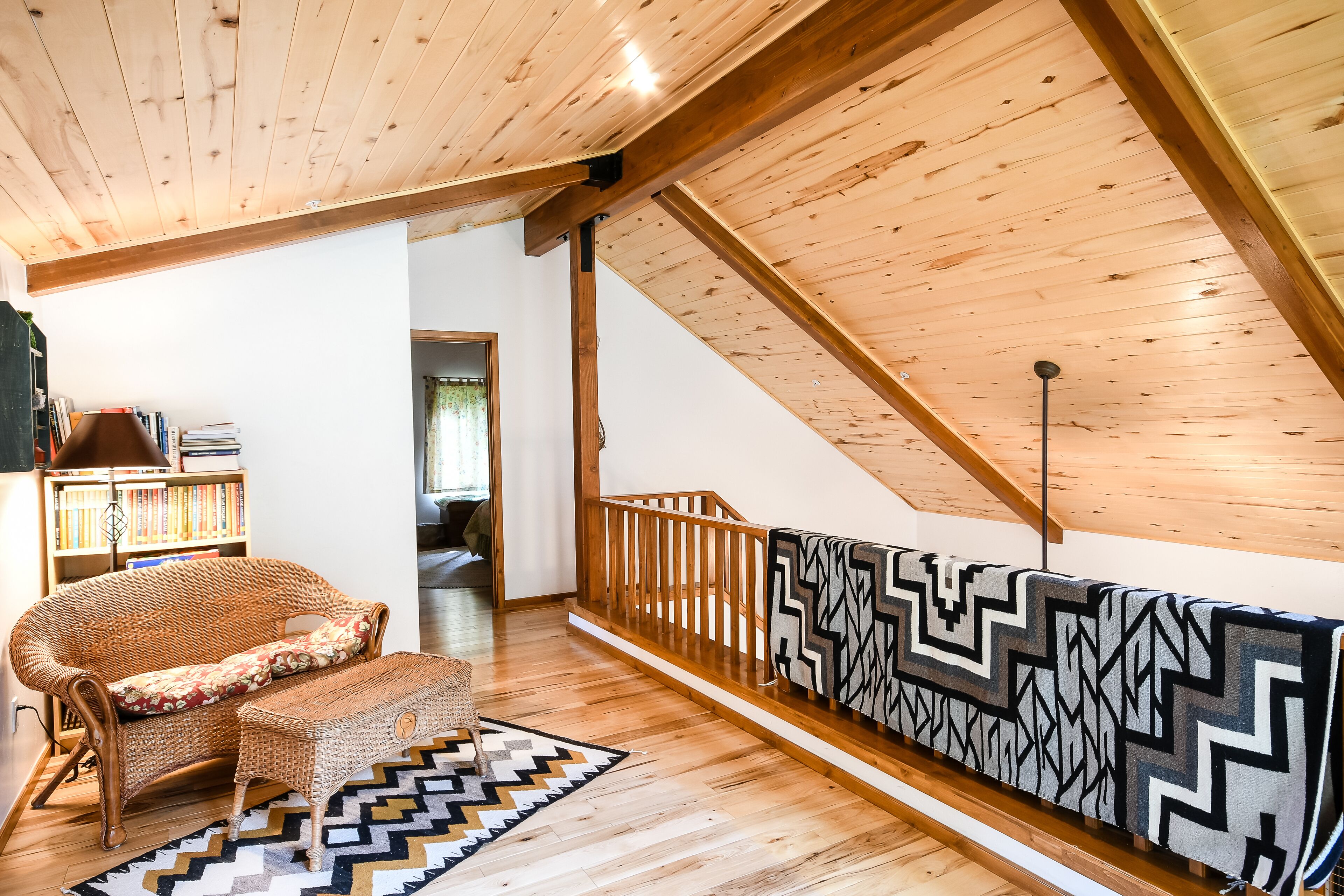 Un loft spacieux avec un plafond haut, des poutres apparentes, un canapé en rotin, un tapis à motifs et une bibliothèque.